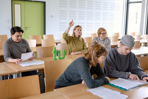 Students in the seminar room are sitting at the tables. One of them is giving a hand signal.